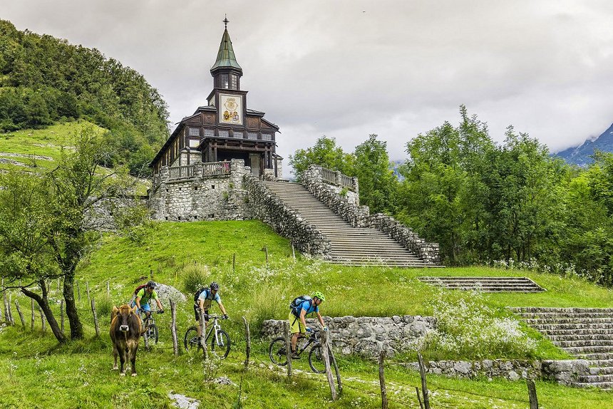 Drei Mountainbiker steigen entlang des Weges unter der Holzkirche in Javorca ab, wo auch eine Kuh weidet.